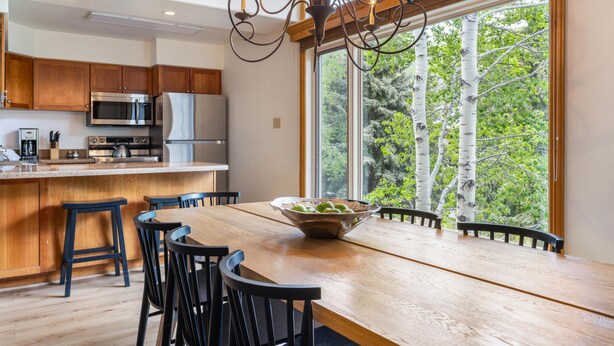 A modern kitchen with wooden cabinets, stainless steel appliances, a large dining table with chairs, and a view of trees through a large window. - Riva Ridge 735 | 4BR Vail Village (Vail)