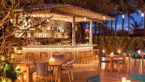 Un área de bar al aire libre con mesas y sillas de madera, rodeada de follaje y palmeras.