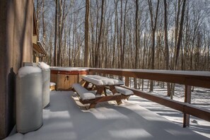 A wooden deck with a hot tub and picnic table covered in snow, surrounded by a forest.