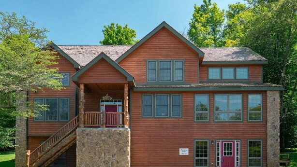 A two-story log cabin with a stone foundation, a porch with a wooden staircase, and a red door.