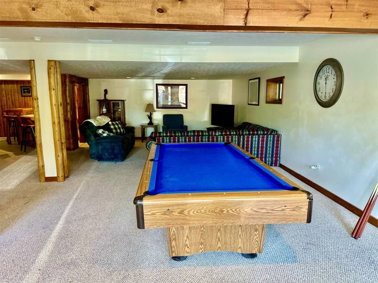 A pool table in a room with a carpeted floor, wooden beams on the ceiling, and a clock on the wall.