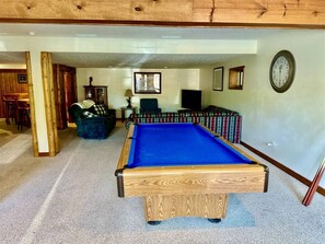 A pool table in a room with a carpeted floor, wooden beams on the ceiling, and a clock on the wall.