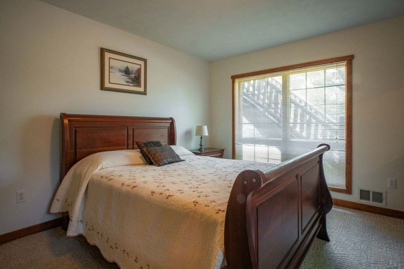 A bedroom with a wooden bed, a nightstand, a lamp, a window with blinds, and a framed picture on the wall.