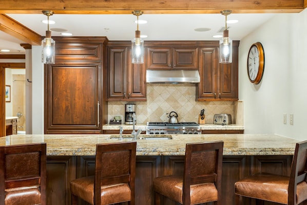 A kitchen with wooden cabinets, a granite countertop, a stainless steel range hood, and a bar area with chairs. - Willows 202 | Luxury 2BR Vail Village (Vail)