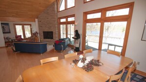 A spacious living room with a large wooden dining table, a fireplace, and large windows offering a view of the outdoors.