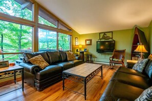 A living room with a leather sofa, a coffee table, and a television. There are large windows providing a view of trees. - Our Greyt Escape - Lake Access and Hot Tub! (Davis)