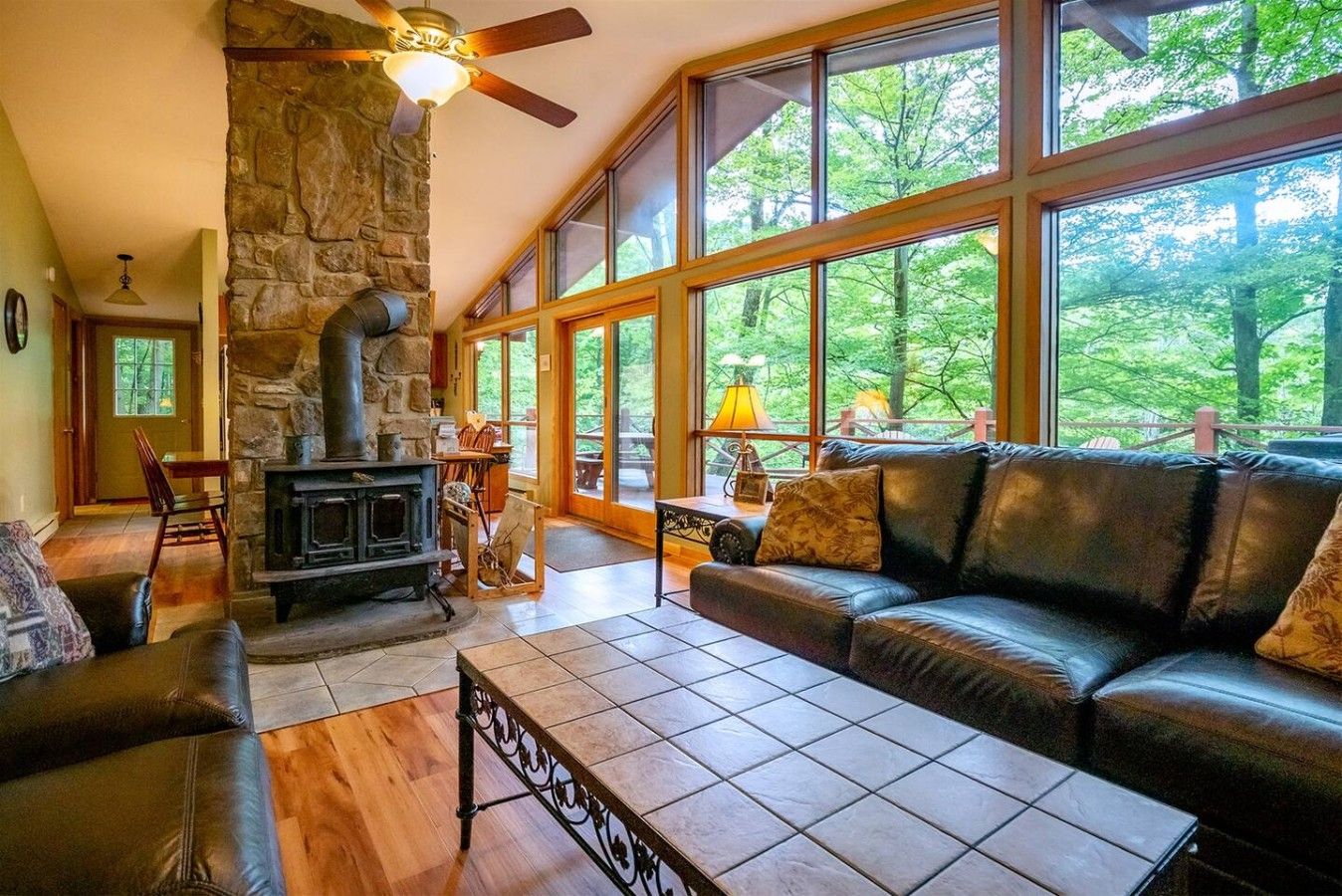 A cozy living room with a stone fireplace, leather sofa, and large windows offering a view of the outdoors.