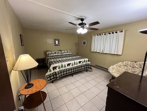 A bedroom with a bed, a ceiling fan, a bedside lamp, a nightstand, and a window with curtains.