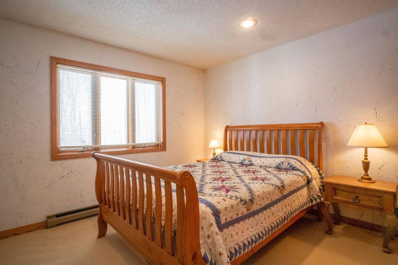 A bedroom with a wooden bed, a window with blinds, a bedside table with a lamp, and a wall-mounted heater.