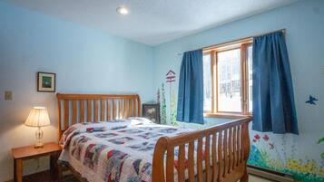 A bedroom with a wooden bed, a bedside table with a lamp, a window with curtains, and a wall mural.