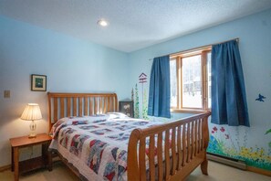 Ein Schlafzimmer mit einem hölzernen Bett, einem Nachttisch mit Lampe, einem Fenster mit Vorhängen und einer Wandmalerei.