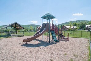 Un parque infantil con un tobogán y estructuras para escalar, rodeado por una valla y casas al fondo.