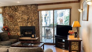 A living room with a stone wall, a fireplace, a sofa, a coffee table, a television, and a balcony with a view of buildings. - Riva Ridge 720 | 2BR Vail Village (Vail)