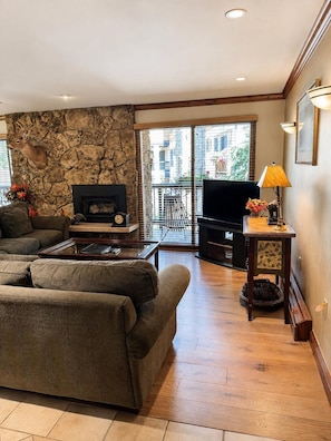 A living room with a stone wall, a fireplace, a sofa, a coffee table, a television, and a balcony with a view of buildings. - Riva Ridge 720 | 2BR Vail Village (Vail)