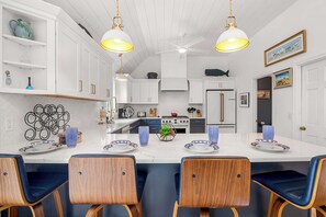 Cocina moderna con armarios blancos, una isla central con sillas azules, iluminación colgante y un comedor con mesa para dos.
