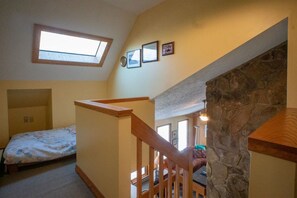 A bedroom with a sloped ceiling, a skylight, a bed, a wooden staircase, and a stone wall.