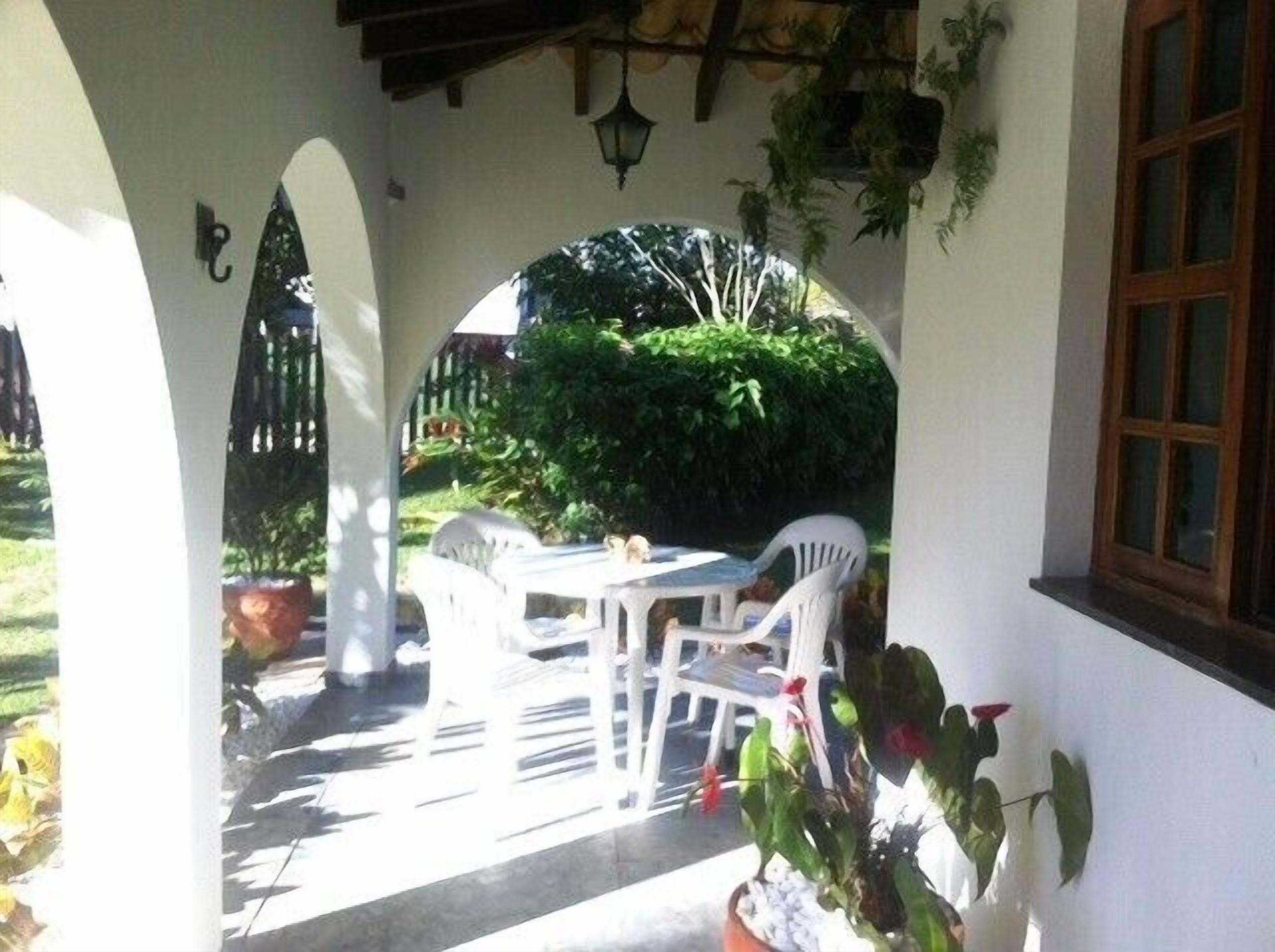 Uma varanda coberta com mesa, cadeiras, plantas em vasos e um marco de janela de madeira.