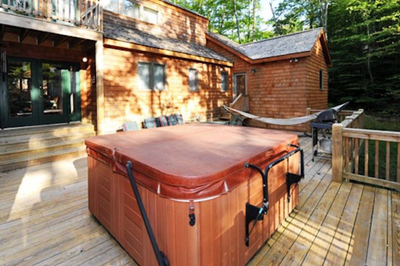 A wooden hot tub on a deck with a hammock and a grill.