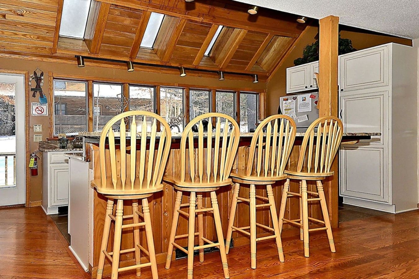 A kitchen with wooden chairs, a white refrigerator, and a window overlooking a snowy landscape.