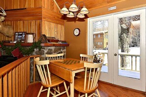 A wooden interior with a dining table, chairs, a clock, and a view through a door.