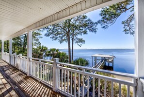 A covered wooden deck with white railings overlooking a calm body of water, a dock, and a small gazebo. - "Bayside Retreat" Panacea FL...Bay Front, Dock, 2 Large Porches, No Pets, Wifi (Panacea)
