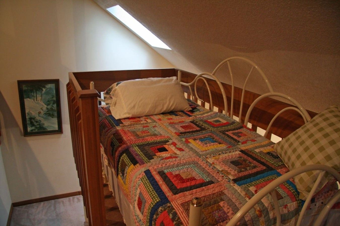 A single bed with a colorful quilt, a white pillow, and a metal headboard in a room with a sloped ceiling and a painting on the wall.