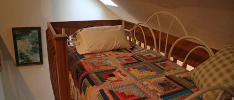 Ein Einzelbett mit einer farbenfrohen Steudecke, einem weißen Kissen und einem Metall-Kopfstück in einem Zimmer mit schrägverdeckter Decke und einem Bild an der Wand.