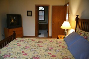 A bedroom with a bed, a television on a dresser, a door leading to a bathroom, and a nightstand with a lamp.