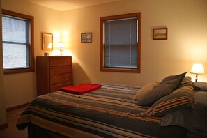 A bedroom with a bed, two bedside lamps, a dresser, and two windows with blinds.