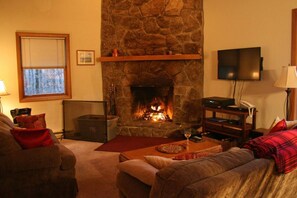 Een gezellige woonkamer met een stenen openhaard, een aan de muur bevestigde televisie en een comfortabele zithoek met een rode plaid.