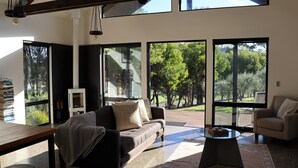 Una sala de estar moderna con un sofá, sillones, una mesa de centro de vidrio y una chimenea. Amplias ventanas ofrecen vistas a un jardín y árboles.