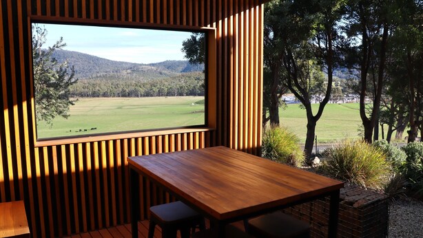 Una mesa y un banco de madera en una habitación con un amplio ventanal que da a un campo cubierto de hierba y árboles.