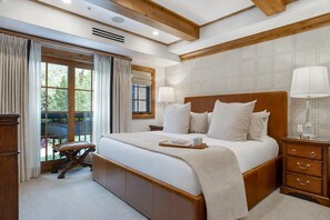 A spacious bedroom with a large bed, wooden headboard, two bedside tables with lamps, a nightstand, and a view of the outdoors through a window.
