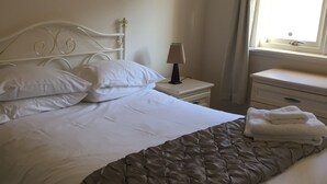 Een net opgemaakt bed met witte lakens en een bruine geborduurde plaid, een bedlampje en een raam met gordijnen.