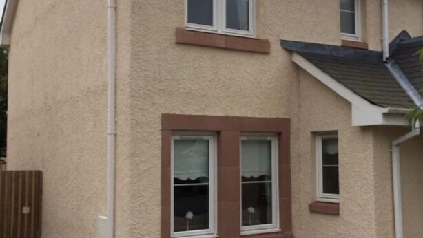 Een twee verdiepingen tellend huis met een beige gevel, witte omlijsting rond ramen en deuren, en een schuin dak.