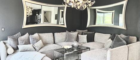 A modern living room with a grey sectional sofa, a glass coffee table, and a large chandelier.