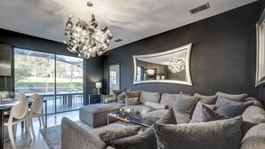 Een moderne woonkamer met een grote grijze bank, een glazen salontafel en een kroonluchter. Er is een eethoek met een tafel en stoelen, en een groot raam met uitzicht op de omgeving.