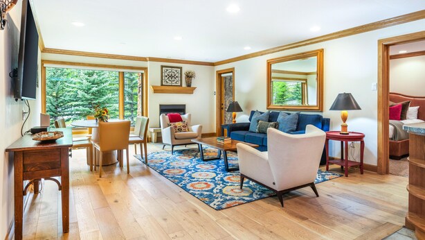 A spacious living room with a wooden floor, a blue patterned rug, a blue sofa, a white armchair, a small table with a lamp, and a fireplace. - Riva Ridge 715 | 2BR Vail Village (Vail)