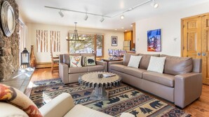 A modern living room with a large sectional sofa, a round coffee table, and a stone wall. There are two windows providing natural light, and a painting is visible on the wall. - Riva Ridge 655 | 2BR Vail Village (Vail)