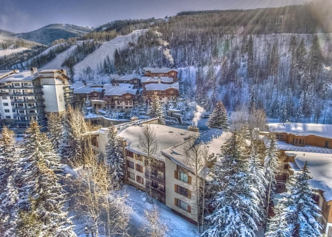 A snow-covered resort with multiple buildings, including a lodge and a hotel, surrounded by trees and a ski slope.