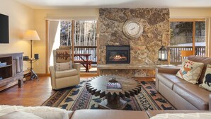 A cozy living room with a stone fireplace, leather sofas, a coffee table, and a large window with a view of trees. - Riva Ridge 655 | 2BR Vail Village (Vail)