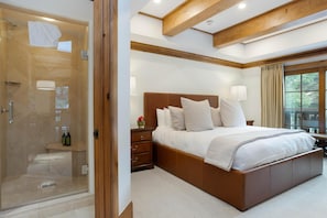 Une chambre à coucher avec un grand lit, une table de chevet, une tête de lit en bois, une cabine de douche en verre et une fenêtre avec des rideaux.