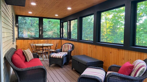 A covered patio with wicker furniture, a round table, and large windows offering a view of greenery. - Quiet, Peaceful Location, Walk to Everything, Hot Tub, Fireplace, Game Room (Lake Placid)
