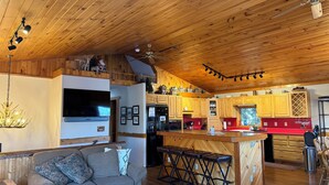 A spacious living room with a wooden ceiling, a sofa, a TV, and a kitchen area with wooden cabinets and a red countertop. - Quiet, Peaceful Location, Walk to Everything, Hot Tub, Fireplace, Game Room (Lake Placid)