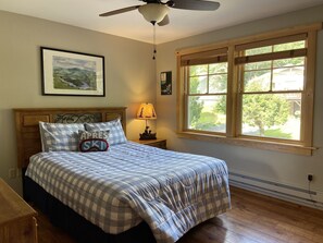 Une chambre à coucher avec un lit, un ventilateur de plafond, une fenêtre avec des stores, une lampe et un tableau encadré au mur.