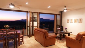 Un balcon offrant une vue sur le coucher de soleil derrière les montagnes, aménagé avec des canapés en cuir, une table à manger et des chaises.