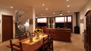Un coin repas avec une table et des chaises en bois, un canapé et une vue sur le coucher de soleil à travers de grandes fenêtres.
