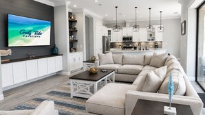 Una sala de estar moderna con un televisor de pantalla plana grande, un sofá modular, una mesa de centro y una cocina al fondo.