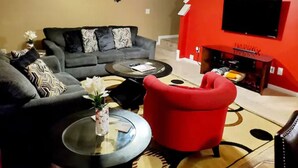 Ein Wohnzimmer mit grauem Sofa, einem roten Sessel, einem Glastisch und einem Kamin, über dem ein Fernseher angebracht ist.
