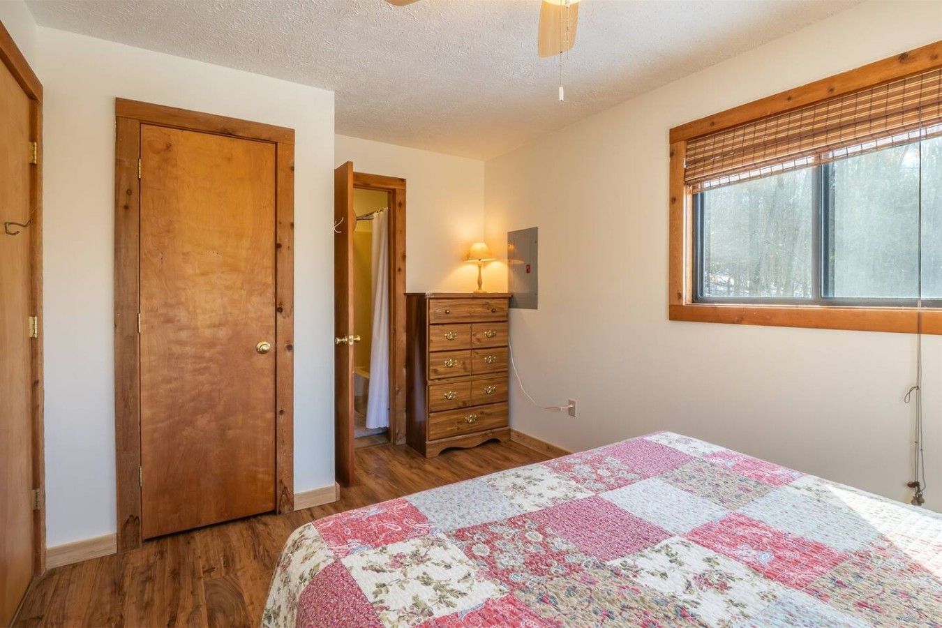 A bedroom with a wooden door, a dresser, a bed with a floral quilt, and a window with blinds.